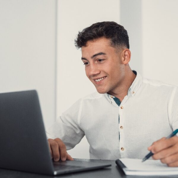 Jovem sorridente usando camisa social branca estuda com um notebook e anotações, representando preparação online para concursos públicos como o do DETRAN RJ.