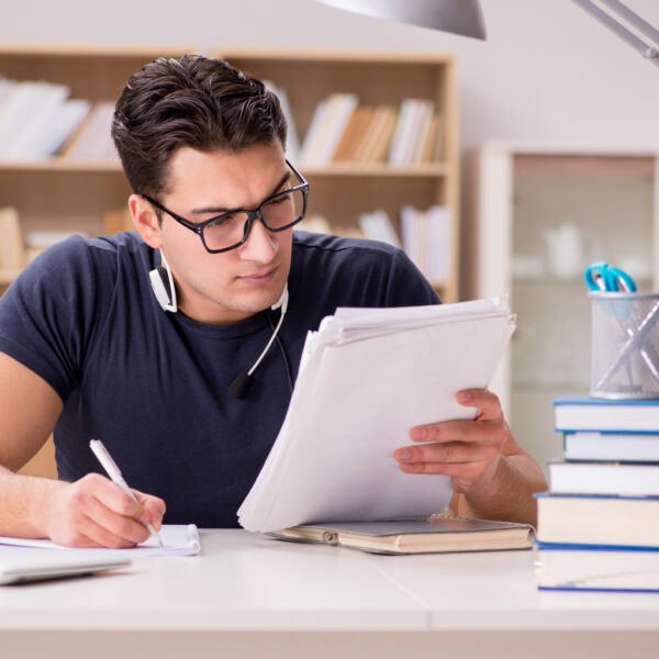Homem jovem estudando com fones de ouvido e livros sobre a mesa, focado na leitura e anotações, representando preparação para concurso público.