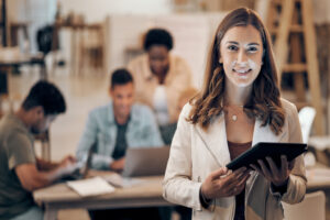 Mulher sorridente com tablet nas mãos em ambiente de trabalho colaborativo, representando uma profissional administrativa pronta para atuar no serviço público.